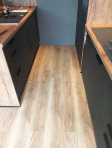 Detail světlé laminátové podlahy Krono Original 6 mm pod tmavými kuchyňskými zásuvkami s dřevěným bokem.